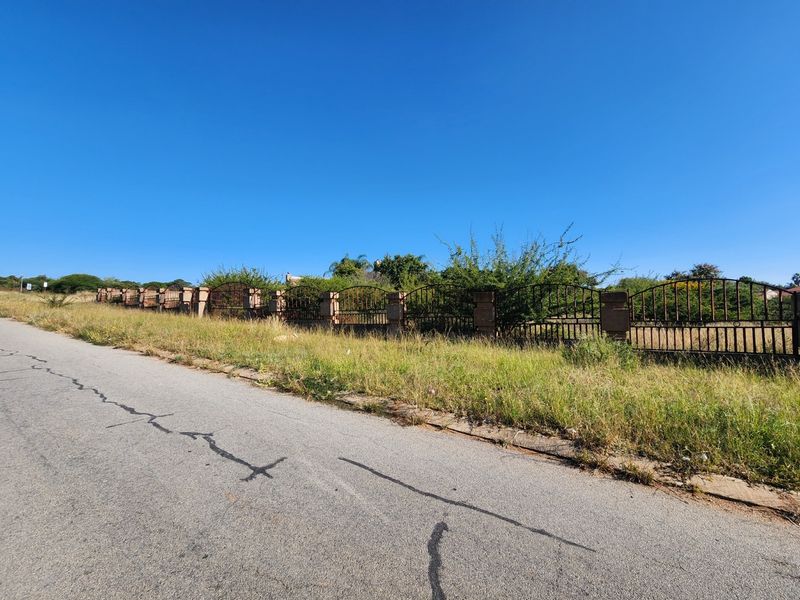 A street-level view of a vacant residential plot featuring a perimeter wrought iron fence with masonry pillars along an asphalt road.