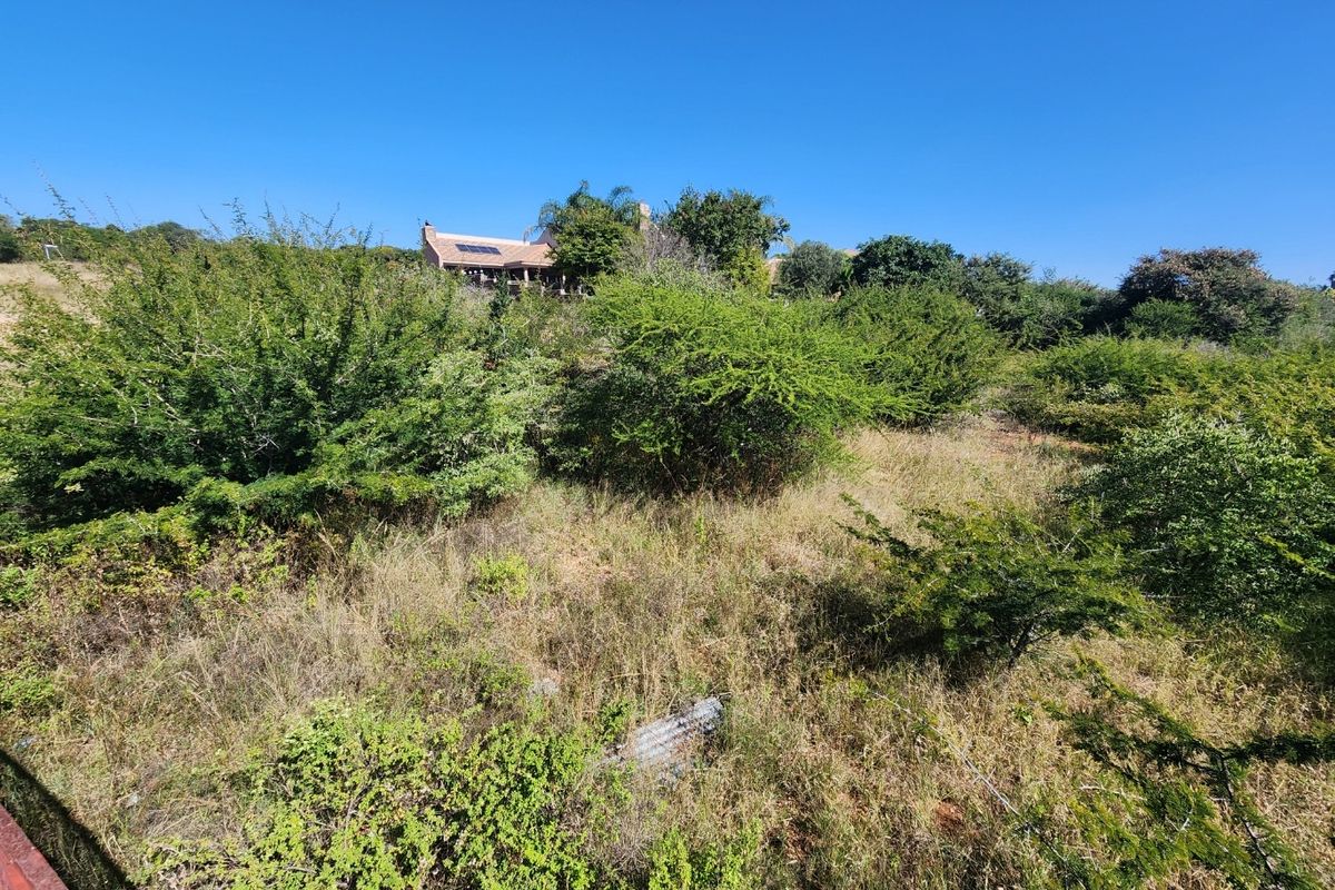 A view of a sloped, undeveloped residential plot featuring natural vegetation and shrubs, with a distant house visible in the background.