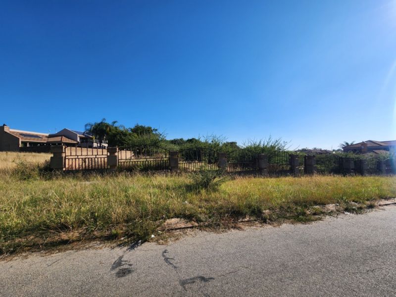 A vacant residential plot featuring a perimeter wrought iron fence with masonry pillars, situated along a paved road in a suburban area.
