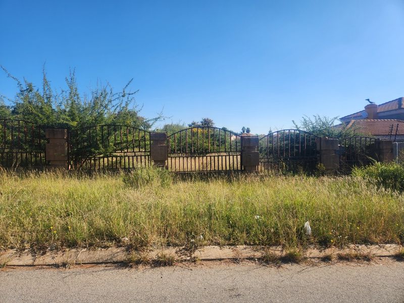A view of a vacant residential plot featuring a wrought iron fence with masonry pillars, bordered by a concrete curb and overgrown grass.