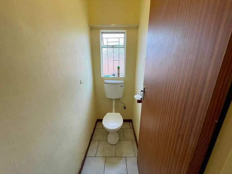 A compact, functional guest toilet featuring tiled flooring and a small window with security bars, requiring cosmetic updates to improve its overall condition.