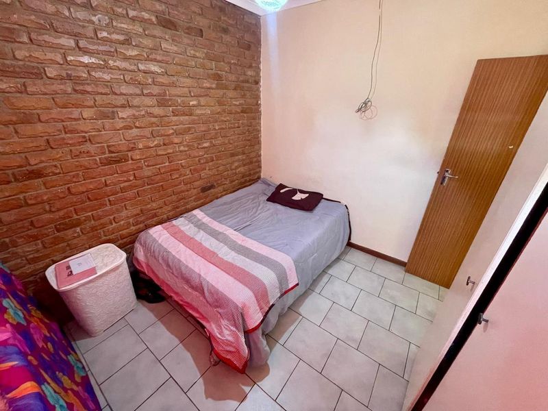 A bedroom featuring an exposed brick feature wall, tiled flooring, and a timber door. The room requires minor cosmetic updates and electrical finishing.