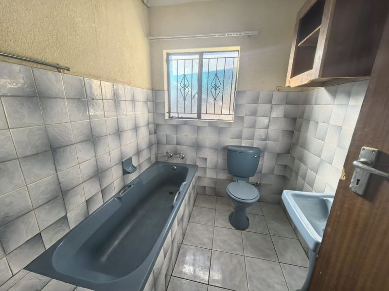 A dated bathroom featuring blue sanitaryware, tiled walls and flooring, and a window with security bars.