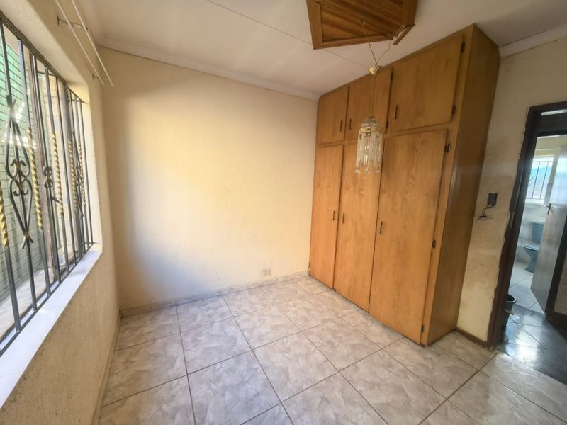 A vacant bedroom featuring built-in wooden cupboards, tiled flooring, and security bars on the window. The room requires cosmetic updates and ceiling repairs.