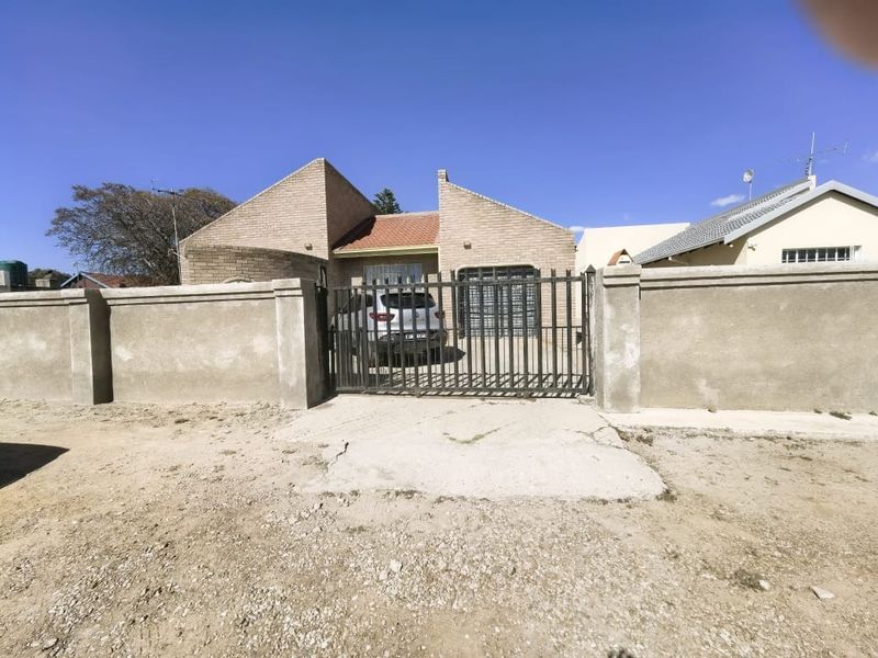 Front view of a brick house featuring a secure metal gate, perimeter wall, and integrated garage, set behind a gravel driveway.