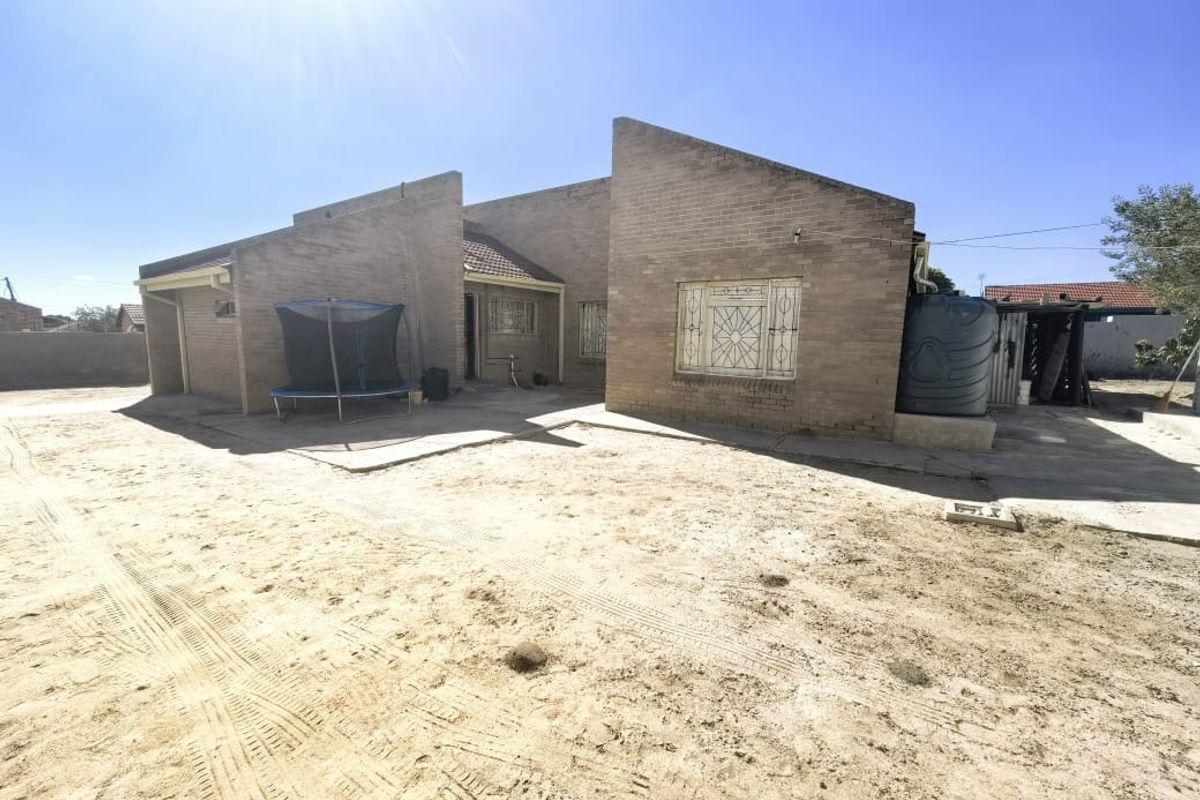 Rear view of a face brick house featuring security bars on windows, a water storage tank, and an expansive unlandscaped sandy yard area.