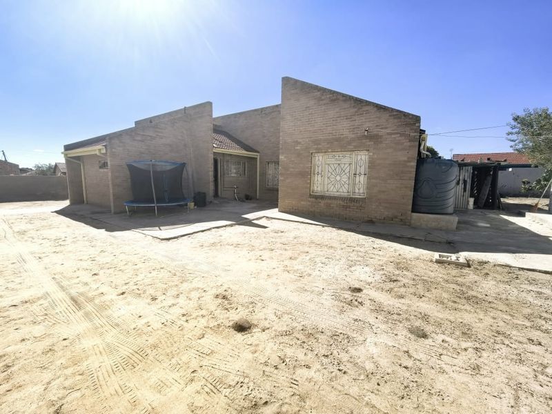 Rear view of a face brick house featuring security bars on windows, a water storage tank, and an expansive unlandscaped sandy yard area.