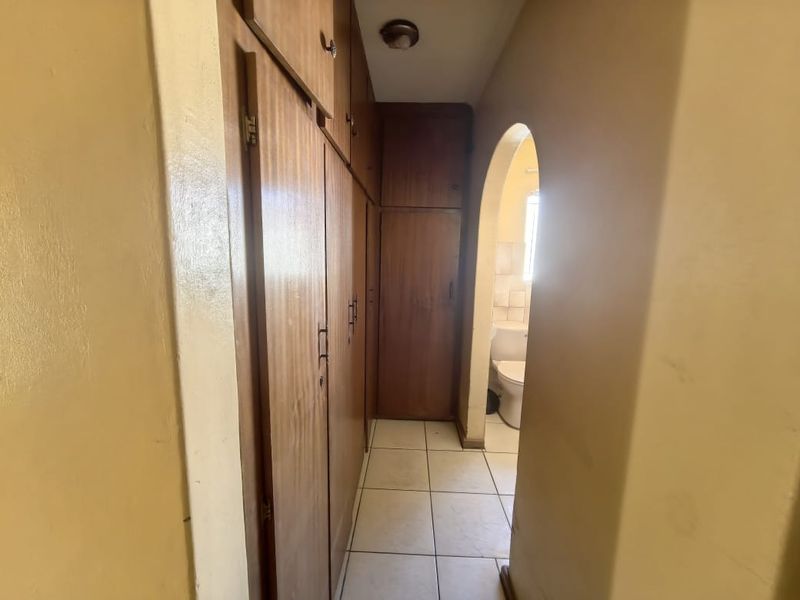 A narrow hallway featuring extensive built-in wooden cabinetry and tiled flooring, leading through an arched doorway into an adjacent room.