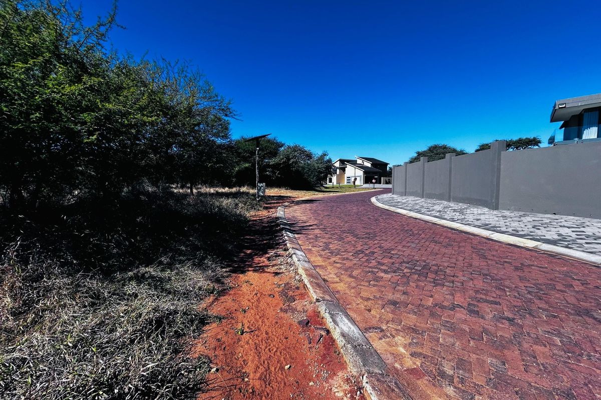 A view of a paved residential street with a concrete boundary wall and established landscaping in a residential estate setting.
