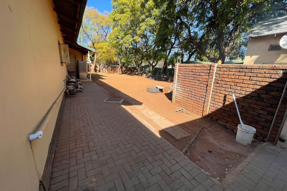 Rear exterior area featuring a paved walkway alongside a brick boundary wall and open garden space with mature trees.