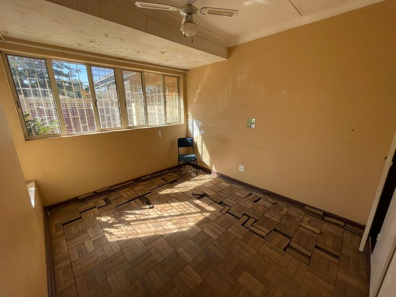 A vacant bedroom featuring parquet flooring, a ceiling fan, and large windows with security bars, currently requiring significant cosmetic repairs and floor restoration.