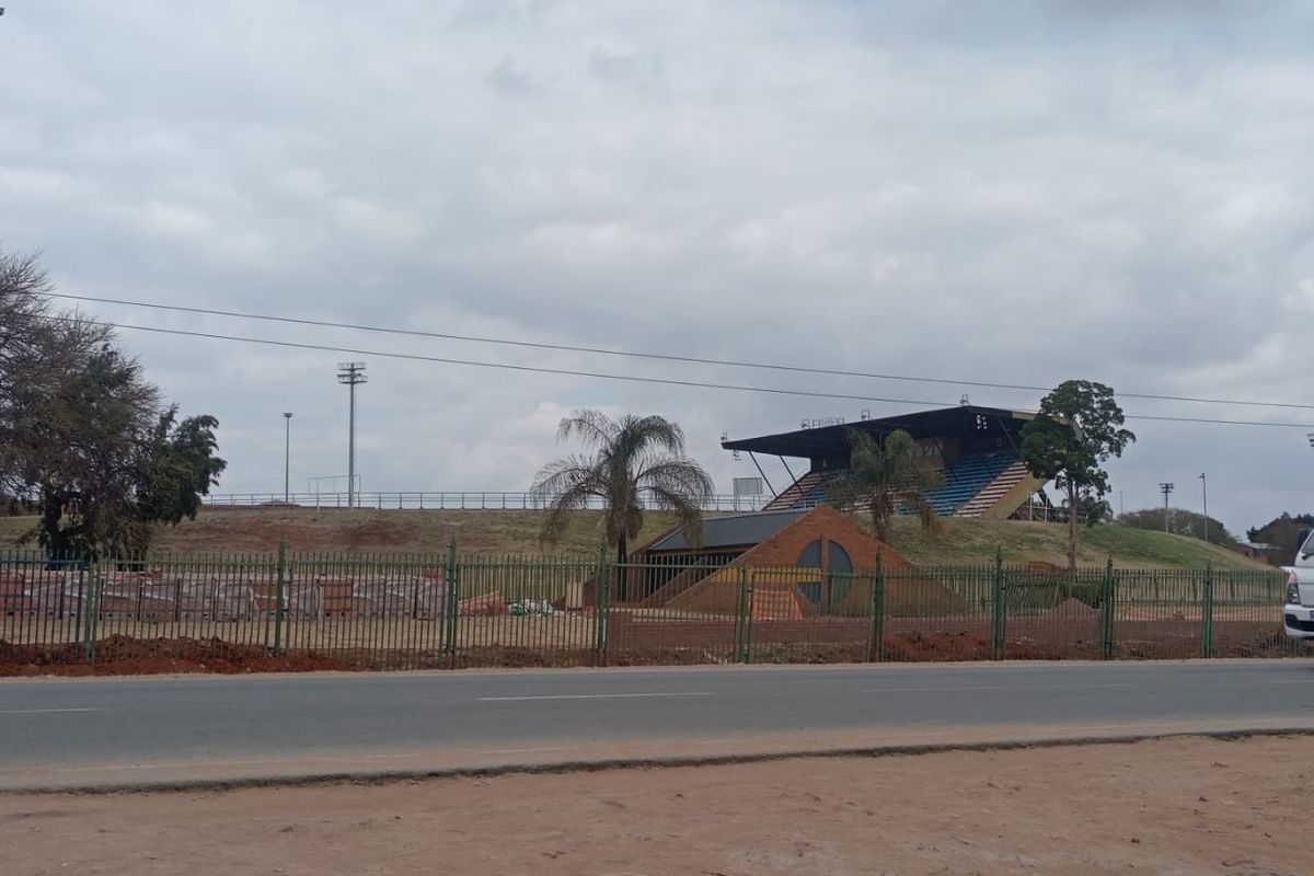 View of a sports facility featuring stadium seating, floodlight masts, and a brick structure, enclosed by perimeter fencing.