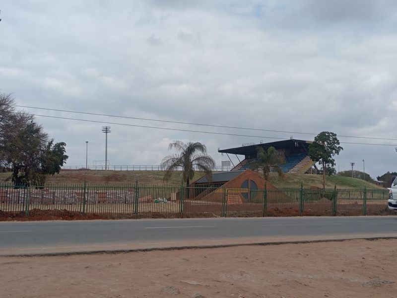View of a sports facility featuring stadium seating, floodlight masts, and a brick structure, enclosed by perimeter fencing.