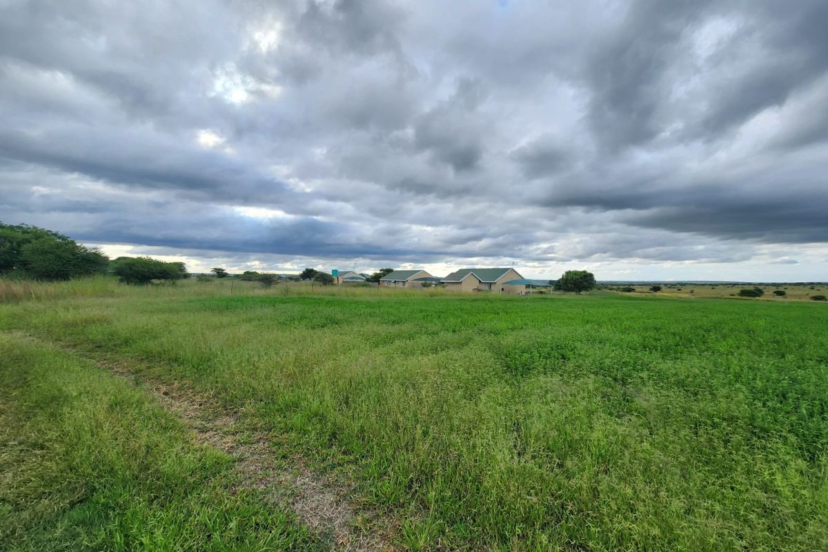 A wide-angle view of the small holding property featuring expansive green fields and distant residential structures under a cloudy sky.