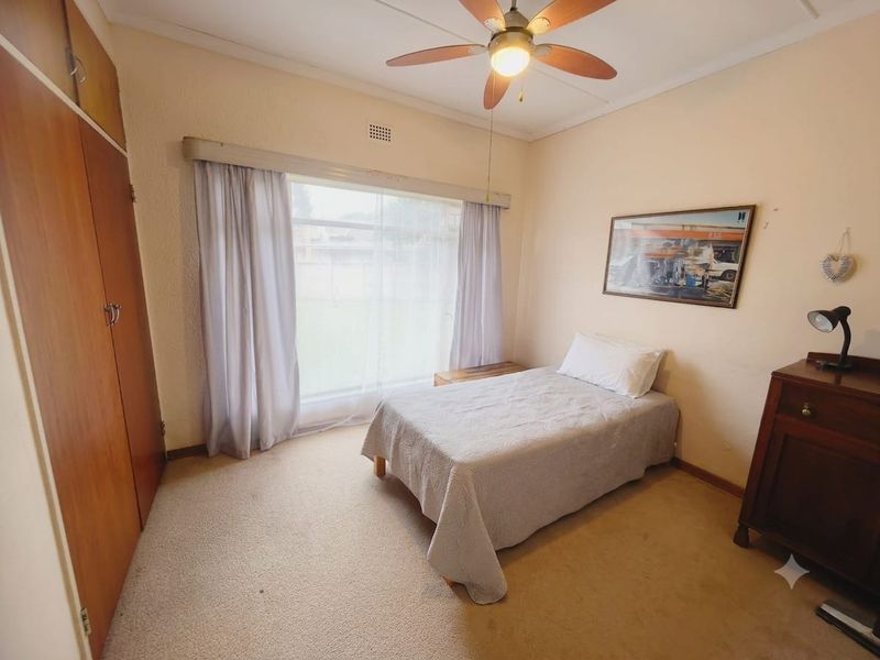 A functional bedroom featuring built-in wooden cupboards, a ceiling fan, and large windows allowing natural light.