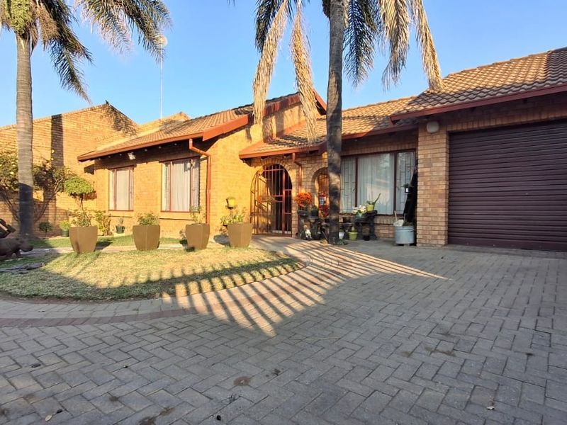 Front view of a face brick townhouse featuring a paved driveway, single garage, and established garden with palm trees.