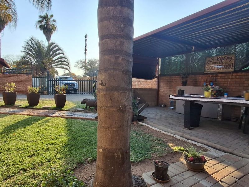 A covered outdoor patio area featuring brick paving and a built-in braai, overlooking a landscaped garden with established trees.