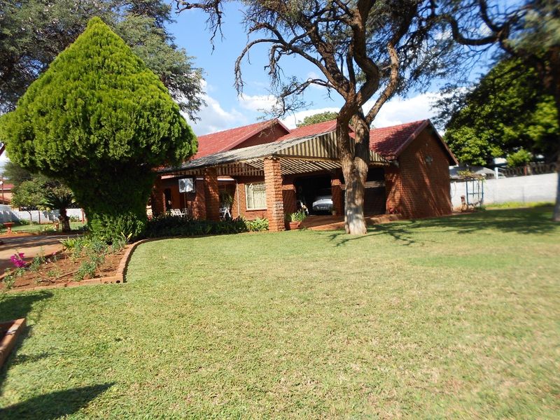 A charming single-story brick house featuring a tiled roof, a spacious covered patio area, and a well-maintained front garden with mature trees.