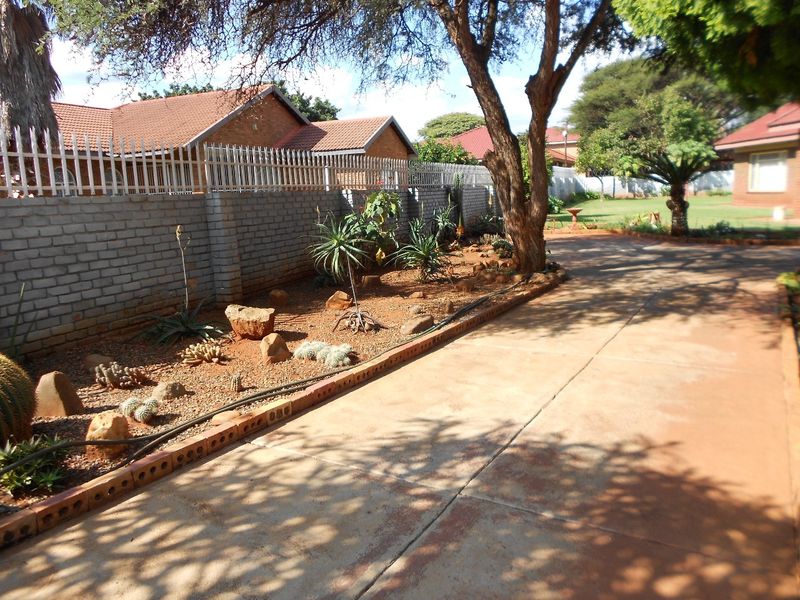 A landscaped garden area featuring a brick boundary wall with security spikes, mature trees, and a concrete driveway with brick edging.