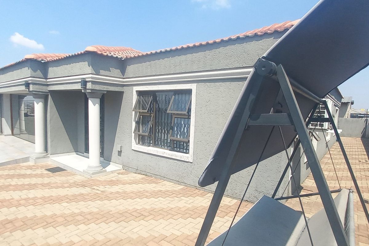 Rear exterior view of the house featuring a paved patio area, textured facade, concrete columns, and secure perimeter fencing.