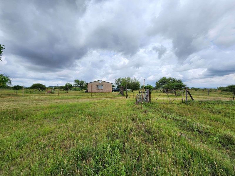 A wide-angle view of a small holding property featuring a detached brick dwelling, wire fencing, and a metal entrance gate set in open land.