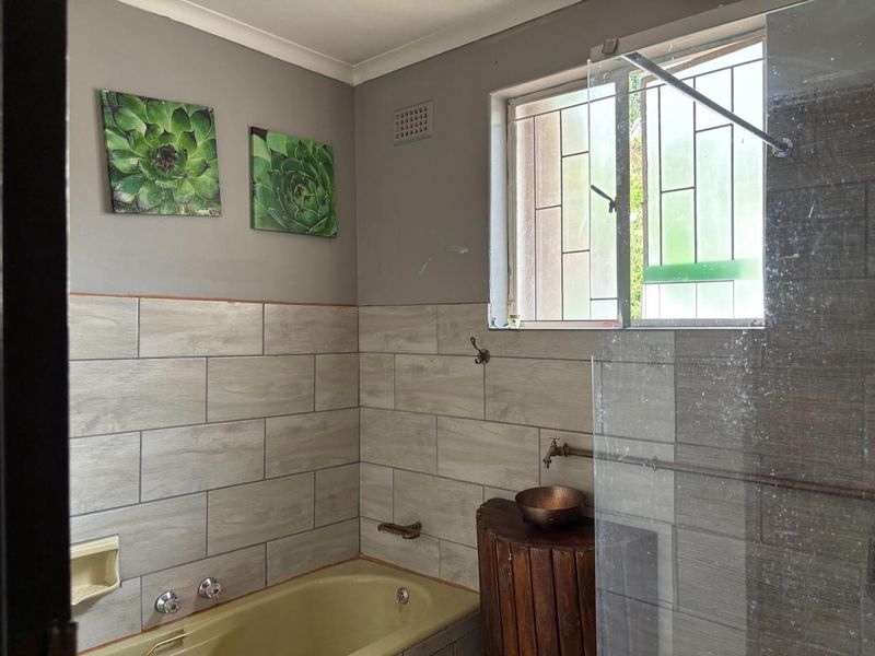 A bathroom featuring a bathtub with a glass shower screen, neutral wall tiling, and a window with security bars.