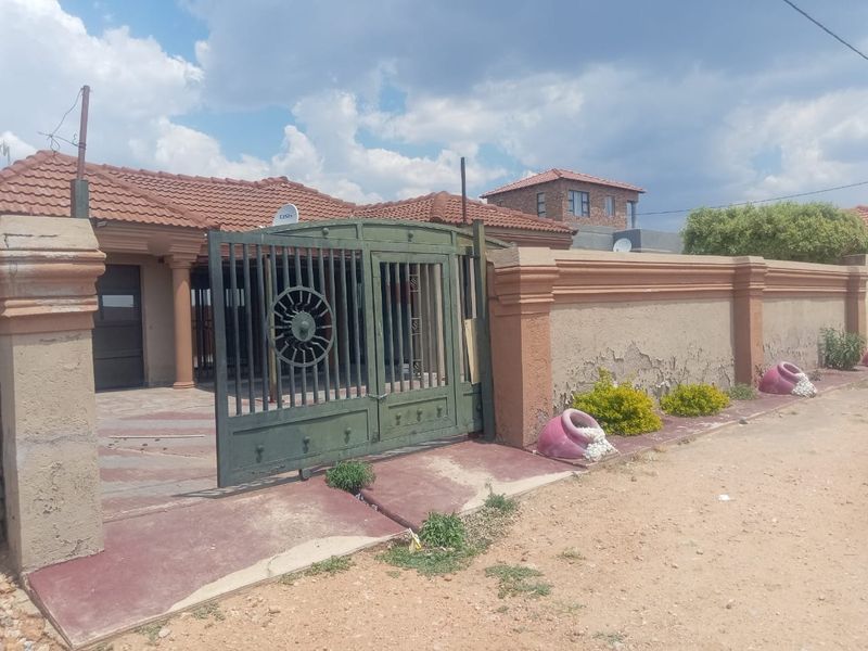 Front exterior view of a house featuring a tiled roof, perimeter wall, and a metal driveway gate with visible wear on the exterior finishes.
