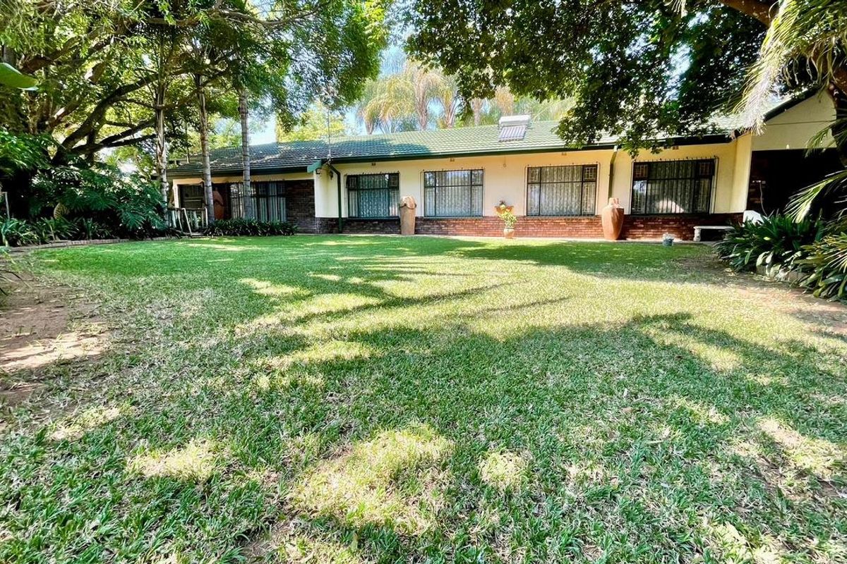 A view of the rear elevation of the house featuring a well-maintained lawn, mature trees, and a brick facade with security bars on windows.