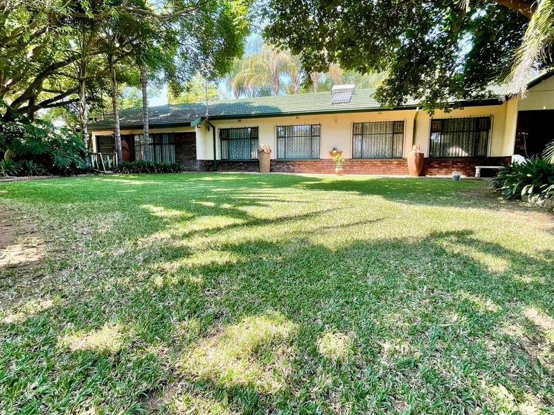 A view of the rear elevation of the house featuring a well-maintained lawn, mature trees, and a brick facade with security bars on windows.