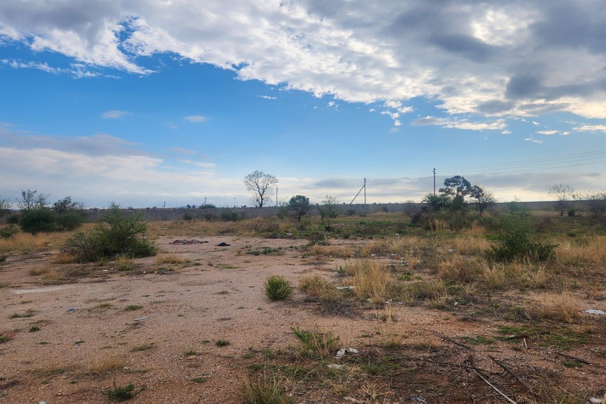 A vacant residential plot featuring natural vegetation, utility poles, and perimeter fencing under a cloudy sky.