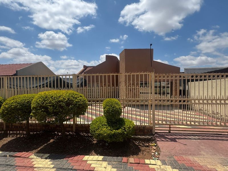 Front view of a residential house featuring a paved driveway, brick facade, and secure metal perimeter fencing.