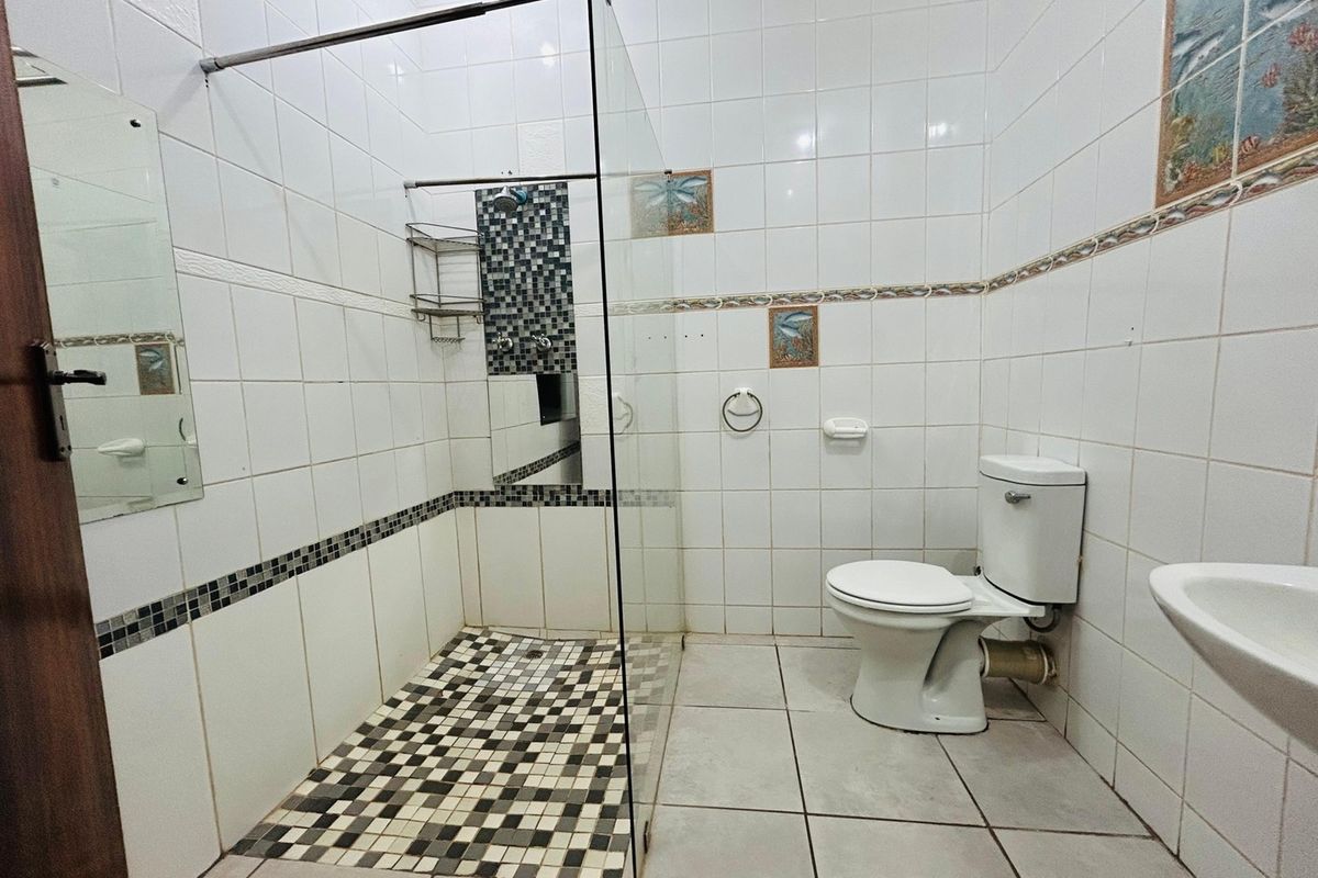 A functional bathroom featuring a glass-enclosed shower with mosaic tiling, a standard ceramic toilet, and a wall-mounted basin with white wall tiles.
