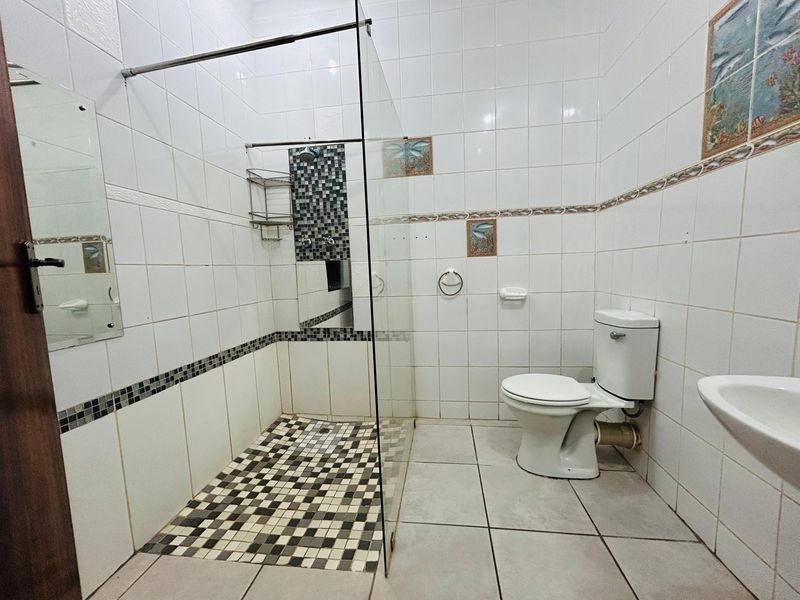 A functional bathroom featuring a glass-enclosed shower with mosaic tiling, a standard ceramic toilet, and a wall-mounted basin with white wall tiles.