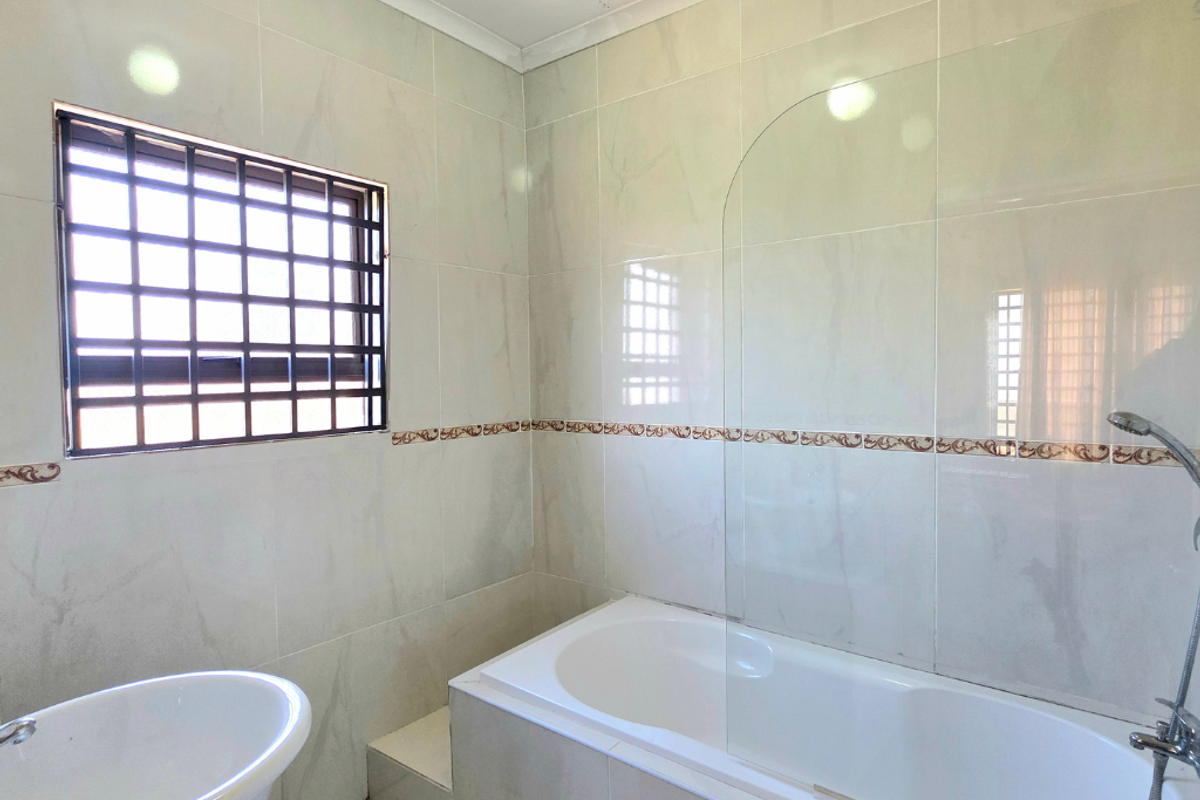 A clean, tiled bathroom featuring a built-in bathtub with a glass shower screen, handheld shower head, and a pedestal basin.