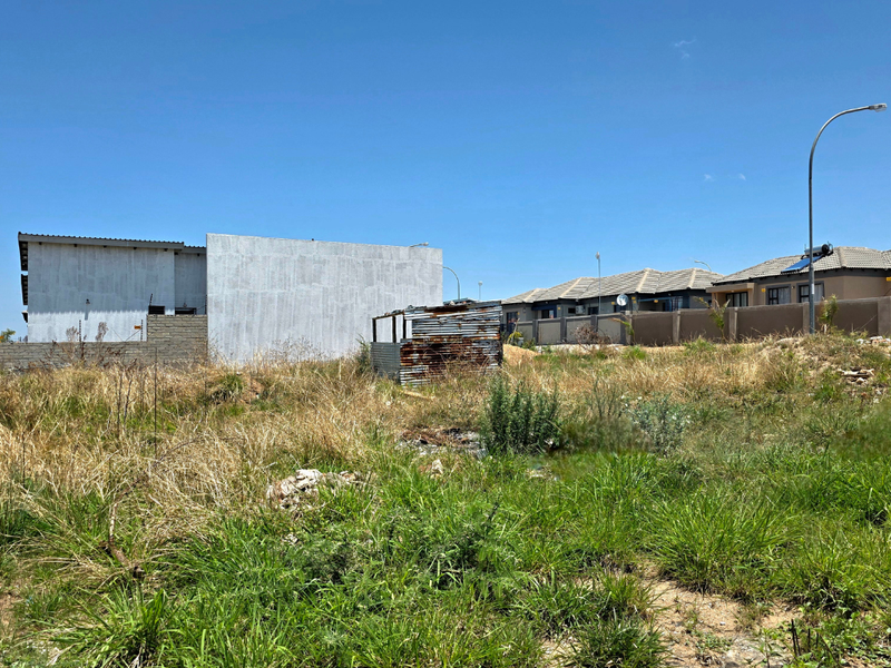 A vacant residential plot featuring overgrown grass, a perimeter wall, and a small dilapidated metal structure, situated in a developed suburban neighborhood.