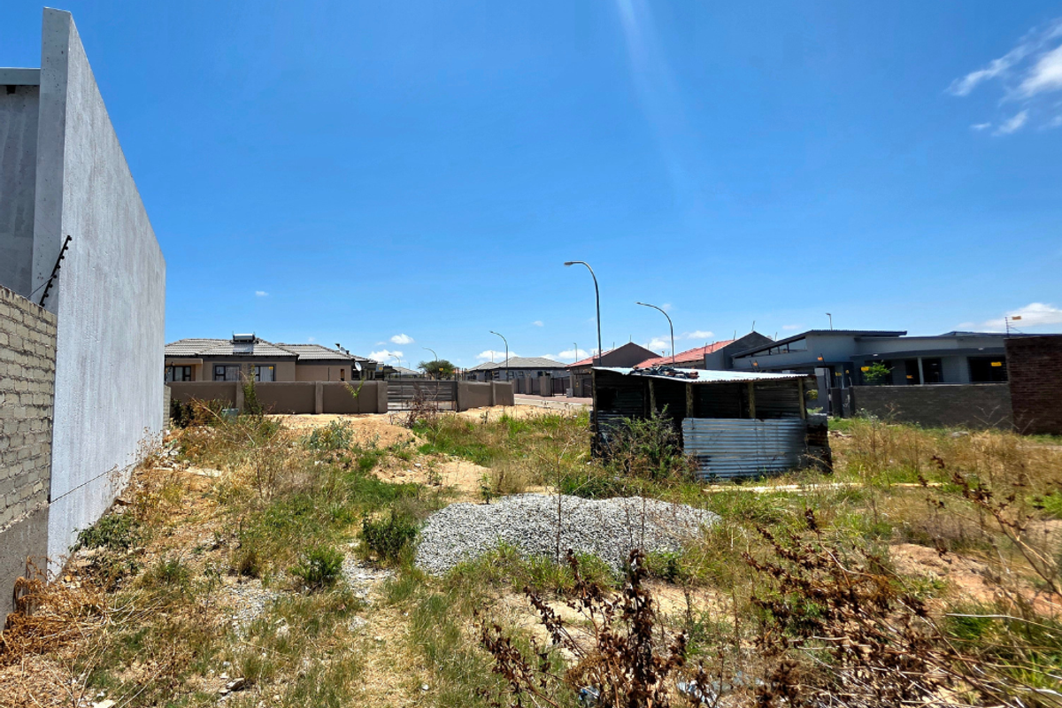 A vacant residential plot featuring a boundary wall with electric fencing, overgrown vegetation, and a small dilapidated temporary structure on site.
