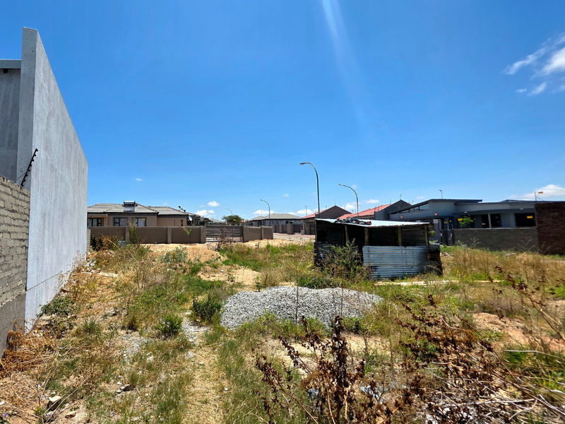 A vacant residential plot featuring a boundary wall with electric fencing, overgrown vegetation, and a small dilapidated temporary structure on site.
