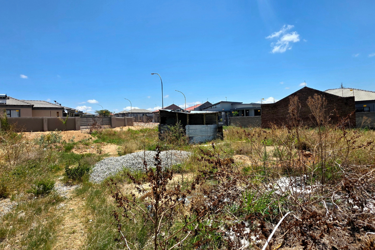 A vacant residential plot featuring overgrown vegetation, a small corrugated metal structure, and surrounding perimeter walls in a developed neighborhood.