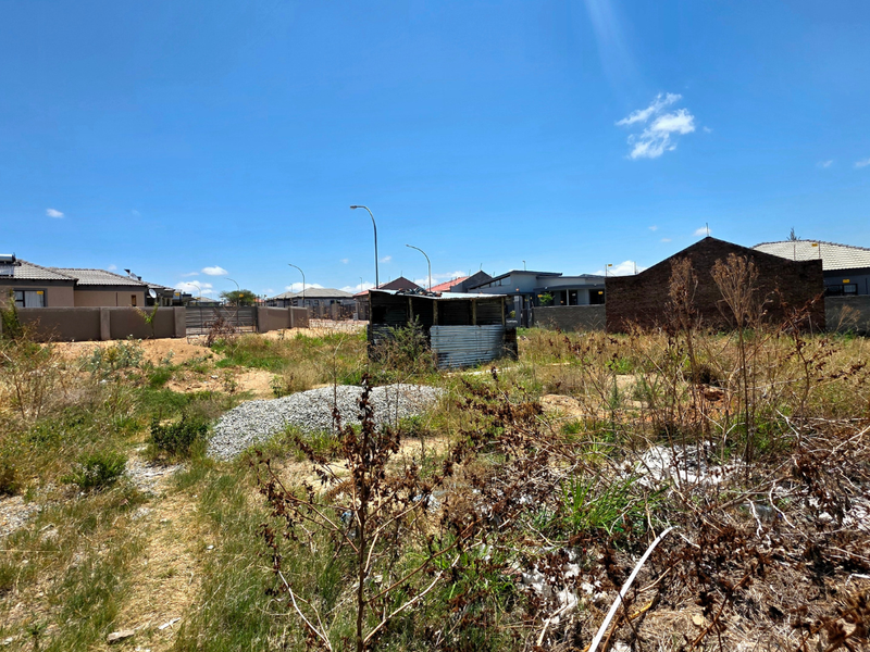 A vacant residential plot featuring overgrown vegetation, a small corrugated metal structure, and surrounding perimeter walls in a developed neighborhood.