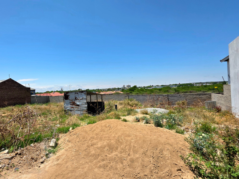 A vacant residential plot featuring a perimeter wall, a small corrugated metal shed, and natural ground cover.