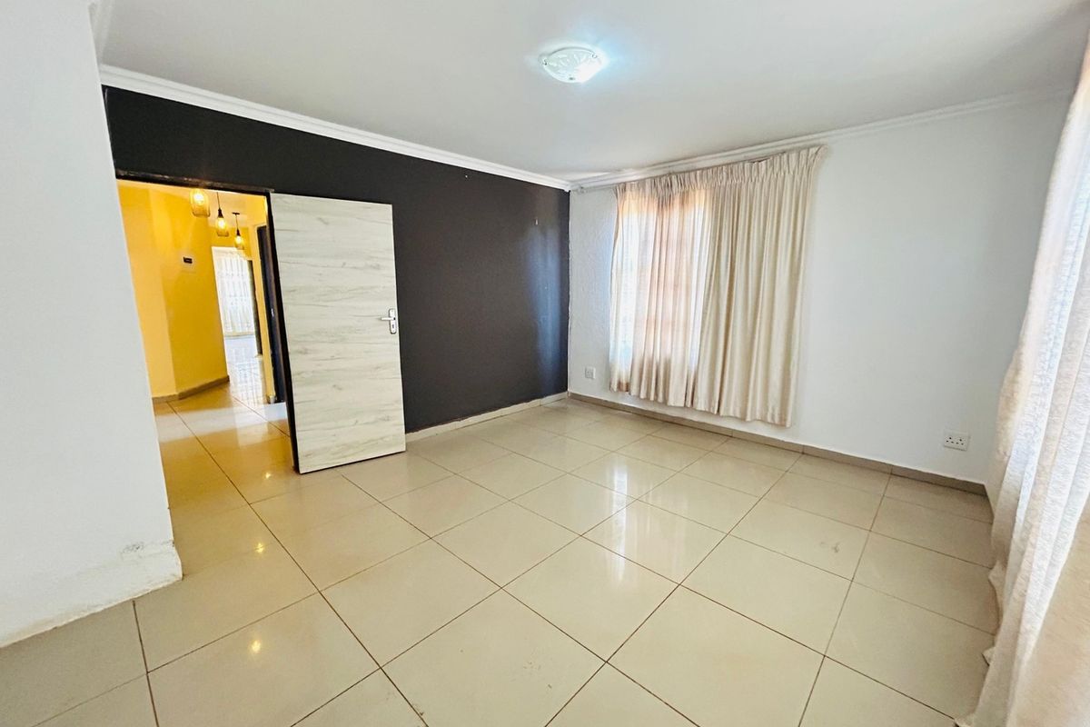 An empty living room featuring neutral tiled flooring, a dark accent wall, and natural light through window curtains.