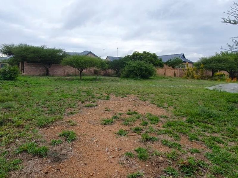 A vacant residential plot featuring established trees, a perimeter brick wall, and a small concrete slab foundation.