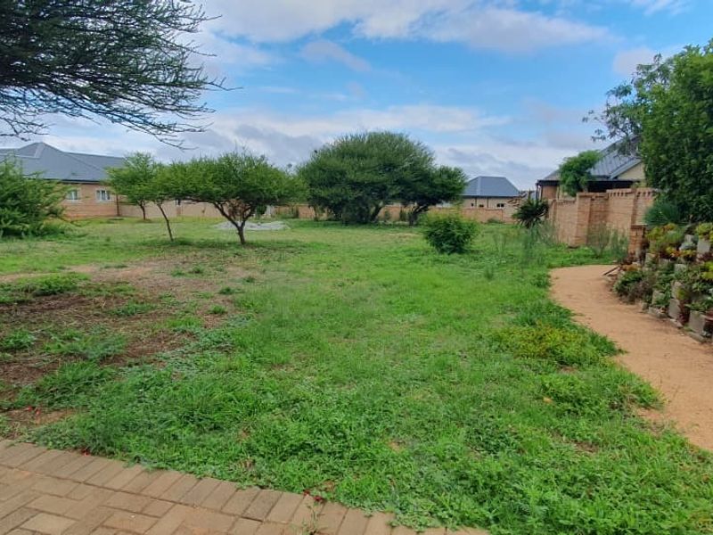 A spacious residential garden featuring a paved patio, a tiered brick retaining wall with established plantings, and a perimeter boundary wall.