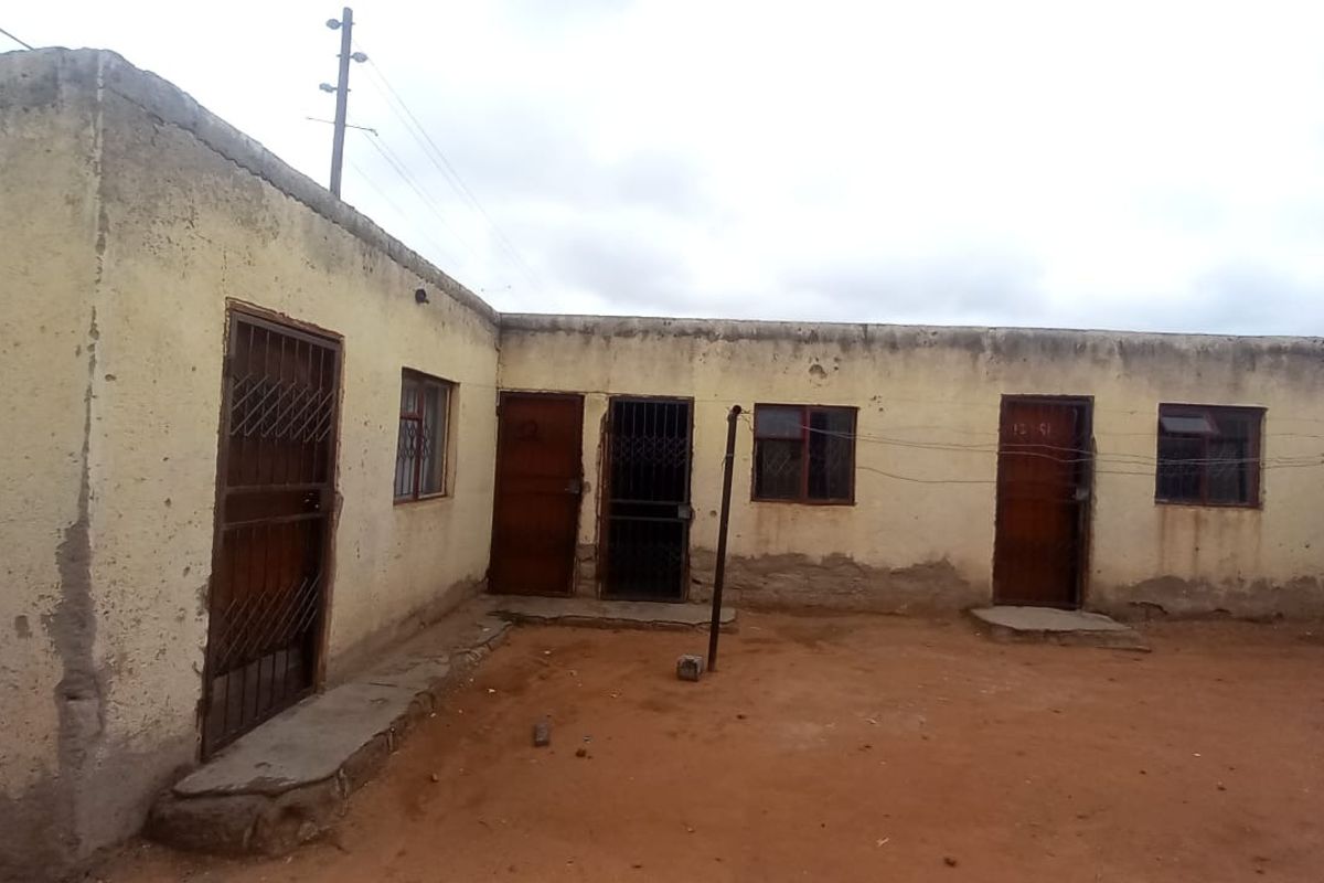 Rear exterior view of a single-story residential building featuring multiple entry points with security gates and weathered plaster walls.