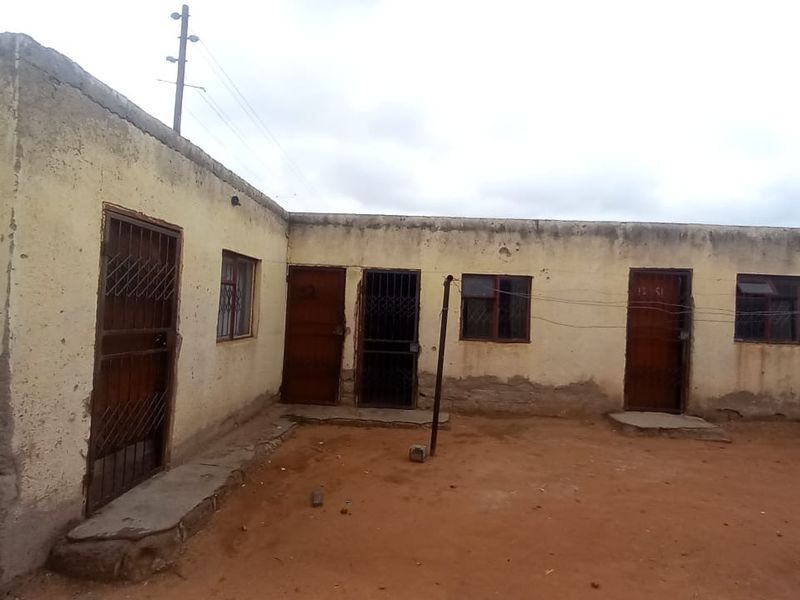 Rear exterior view of a single-story residential building featuring multiple entry points with security gates and weathered plaster walls.