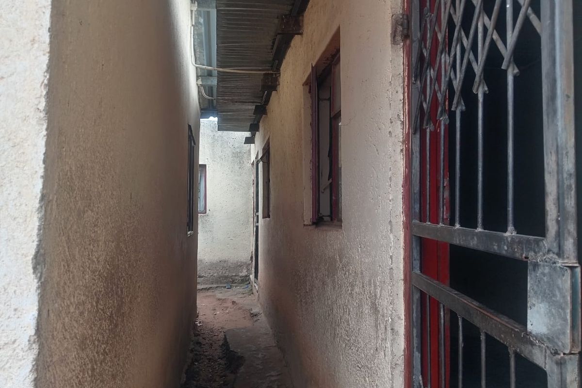 A narrow side passage between building walls featuring security gates and basic window frames with visible wear to the exterior wall finishes.