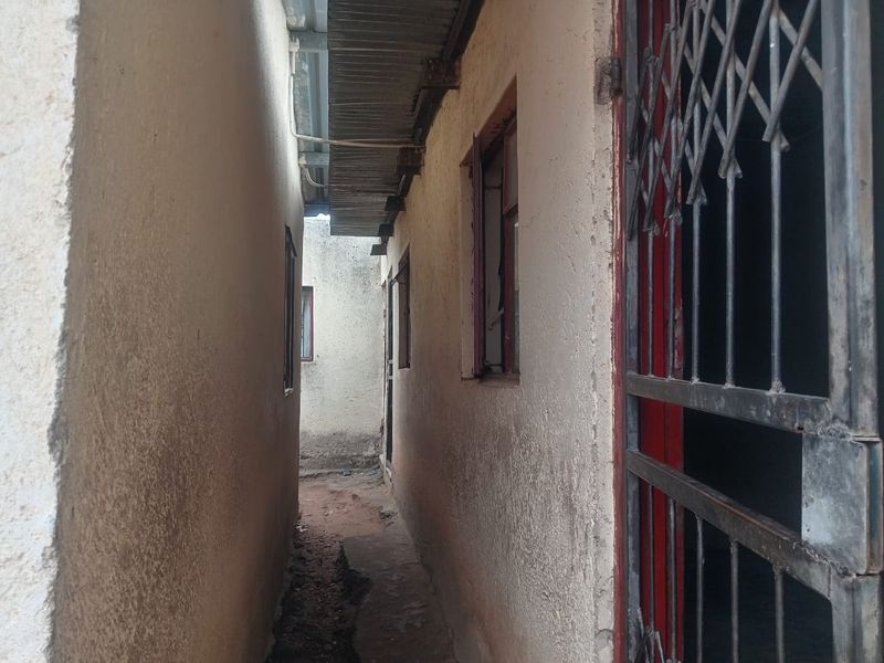 A narrow side passage between building walls featuring security gates and basic window frames with visible wear to the exterior wall finishes.