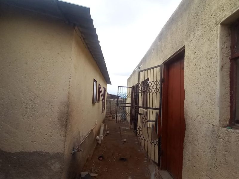 A narrow exterior side passage between two structures featuring textured plaster walls, security gates, and metal window frames with visible signs of wear.