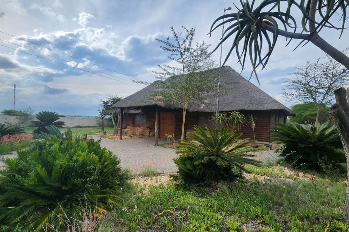 A rustic log cabin with a traditional thatch roof, featuring a paved driveway and established garden surroundings on a small holding property.