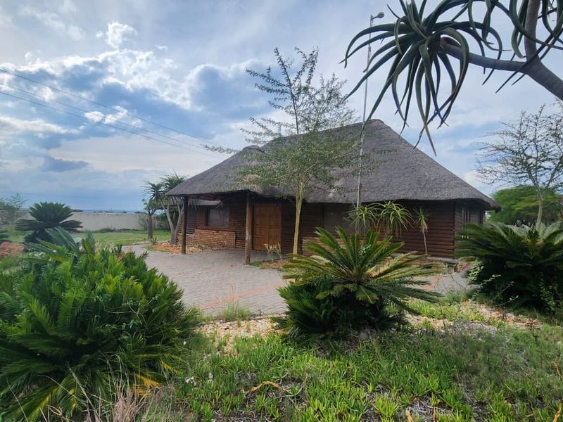 A rustic log cabin with a traditional thatch roof, featuring a paved driveway and established garden surroundings on a small holding property.